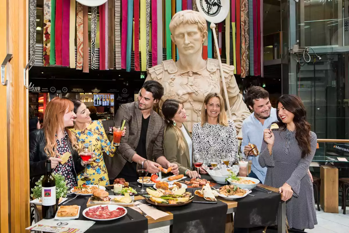 Un grupo de personas disfruta de una comida y bebidas en un ambiente festivo con una gran estatua decorativa al fondo y coloridas cintas colgantes. Un grupo de personas disfruta de una comida y bebidas en un ambiente festivo con una gran estatua decorativa al fondo y coloridas cintas colgantes.