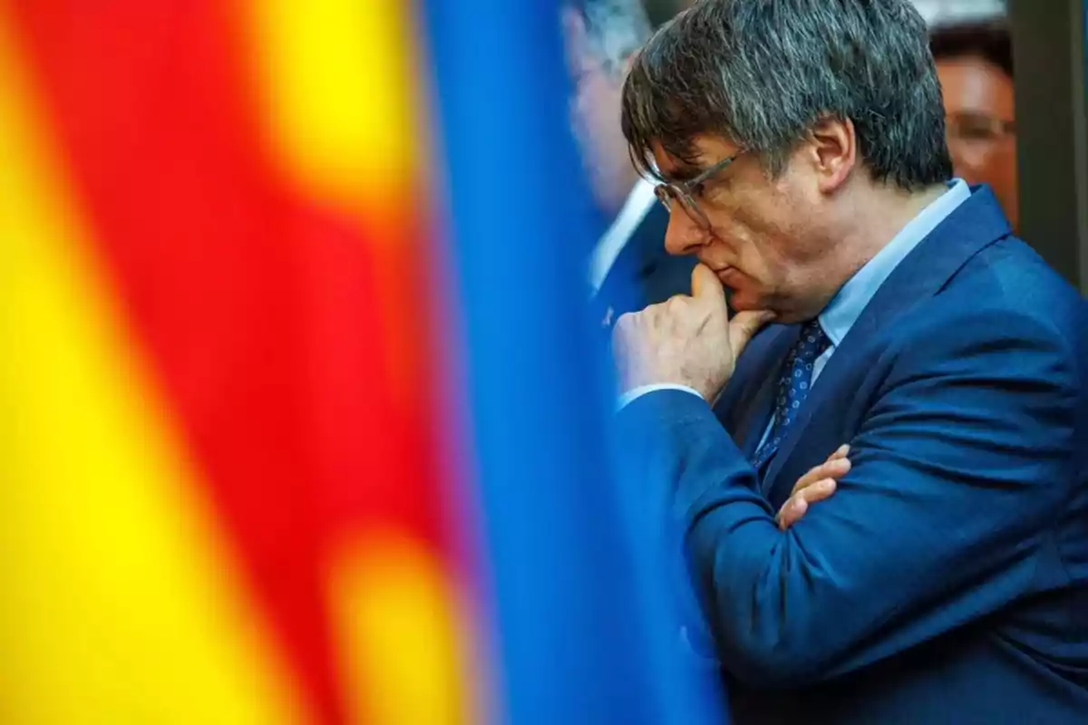 Hombre de perfil con traje azul y gafas, pensativo, con la mano en la barbilla, frente a banderas desenfocadas en primer plano.