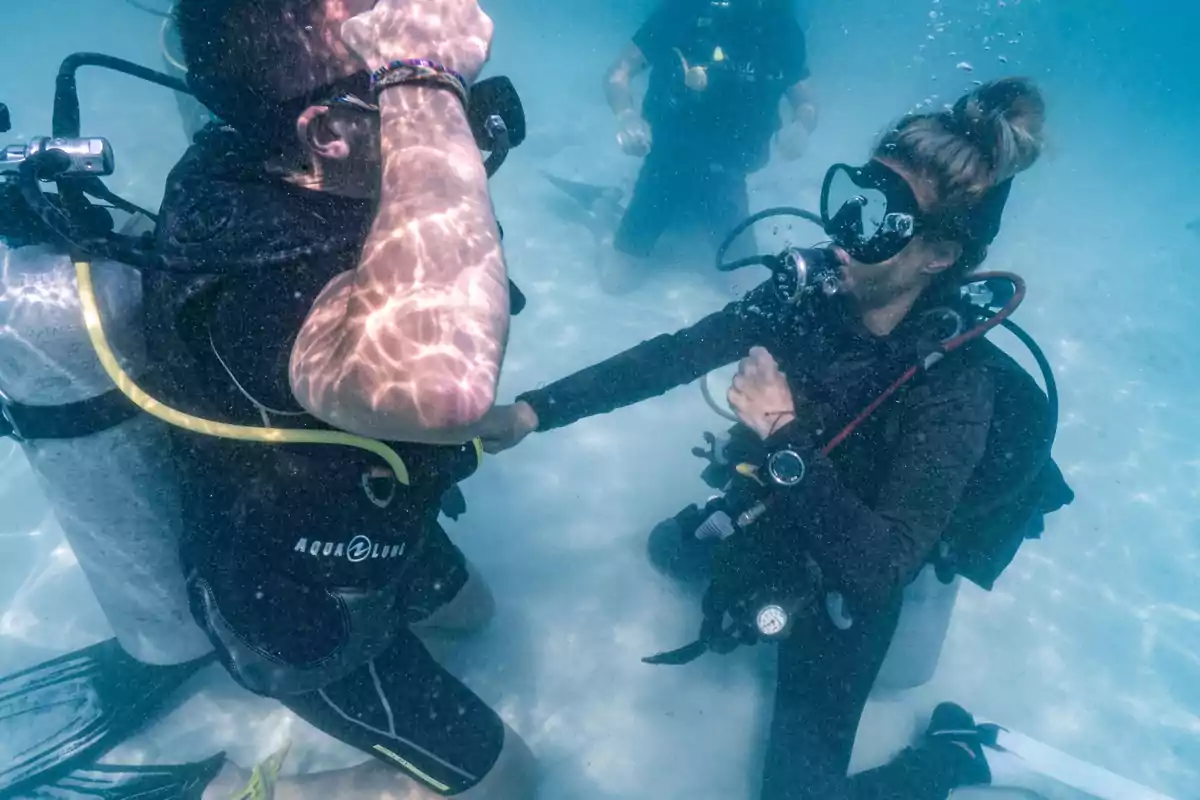 Dos buceadores bajo el agua, uno de ellos parece estar ayudando al otro mientras ambos llevan equipo de buceo.