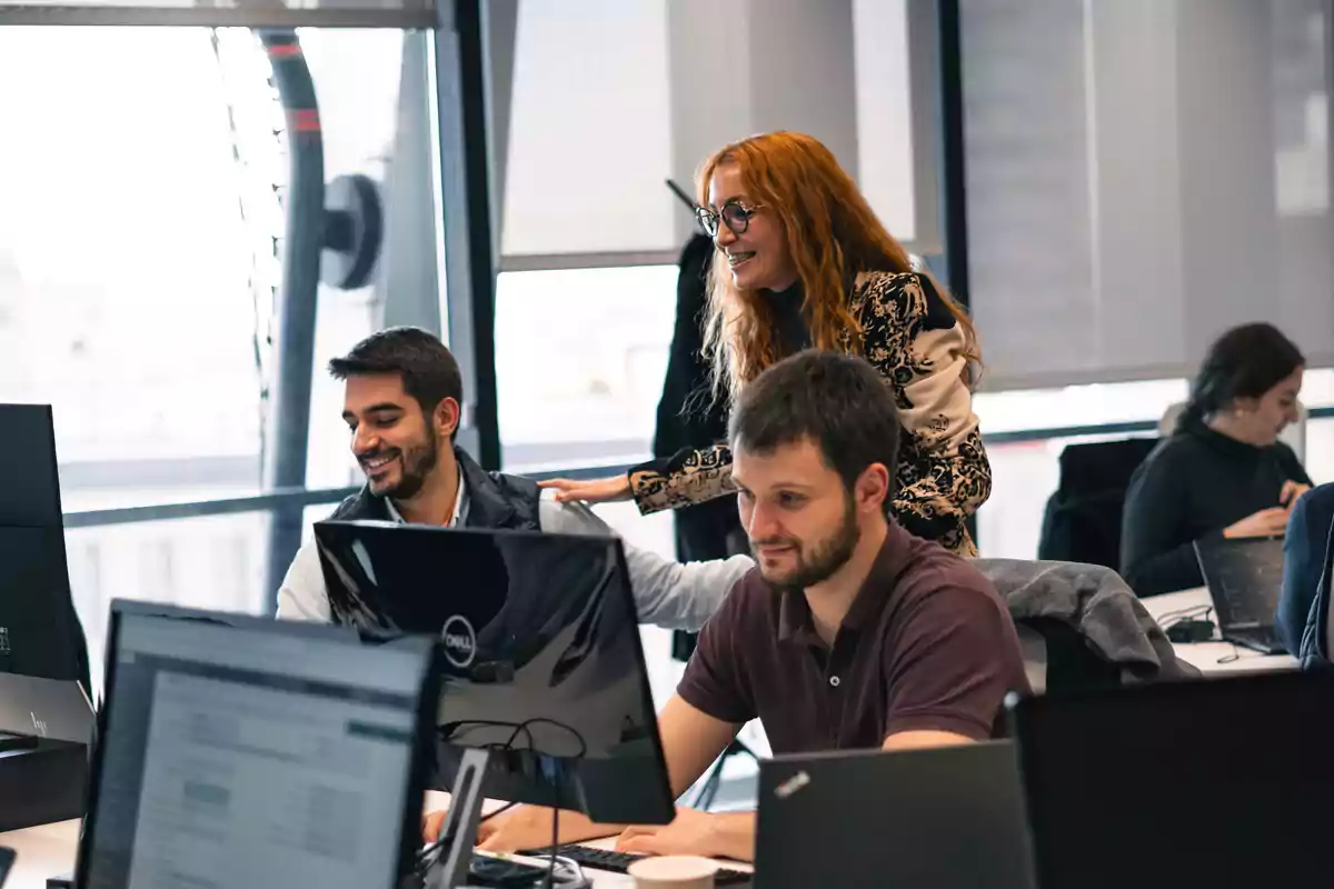 Personas trabajando en una oficina con computadoras y una mujer interactuando con un compañero.