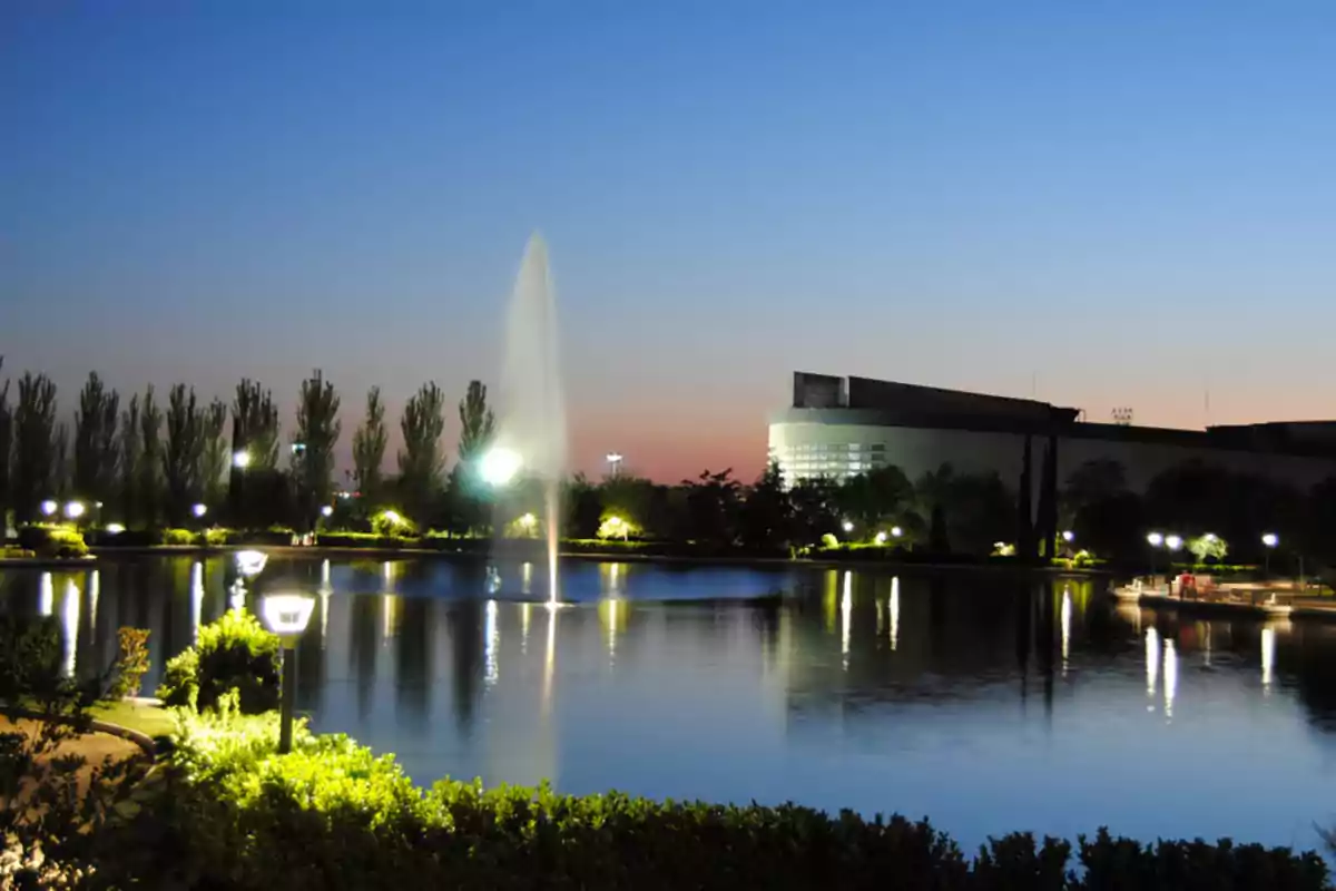 Vista nocturna de un parque con una fuente iluminada en el centro de un lago y un edificio moderno al fondo.