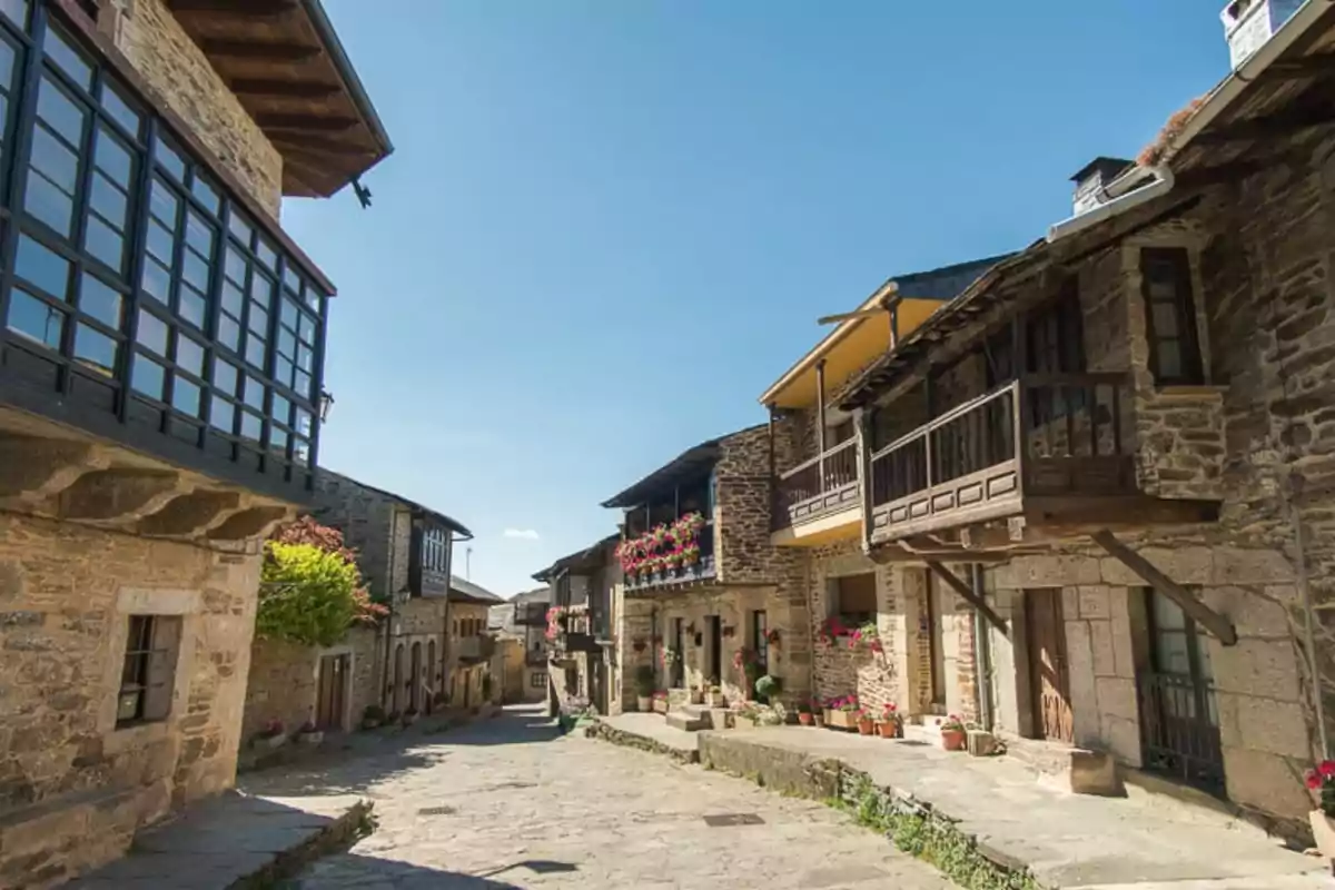 Calle empedrada de un pueblo con casas de piedra y balcones de madera adornados con flores bajo un cielo despejado.