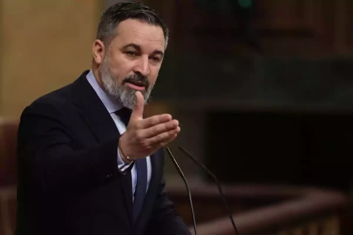 Hombre de barba y cabello corto hablando en un podio con gesto expresivo en un entorno formal