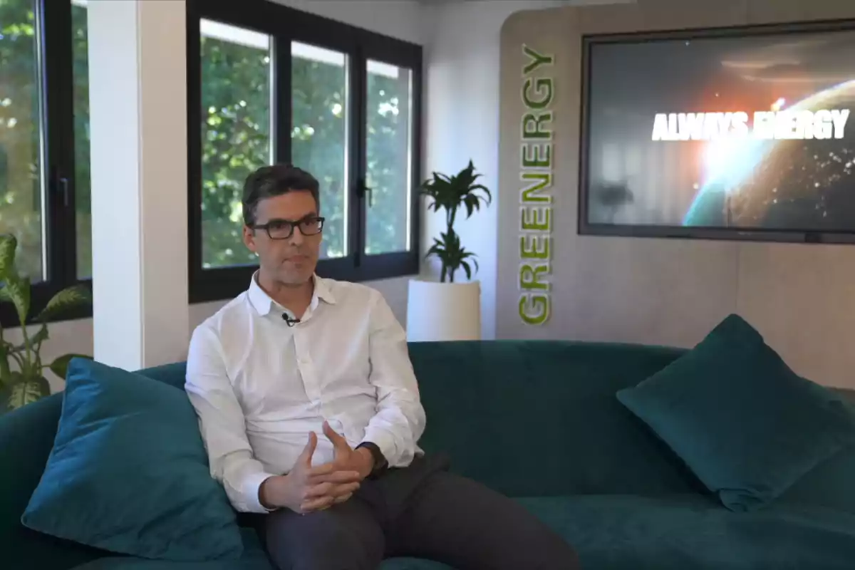 Un hombre con camisa blanca y gafas está sentado en un sofá verde en una oficina moderna con plantas y una pantalla que muestra "Always Energy".