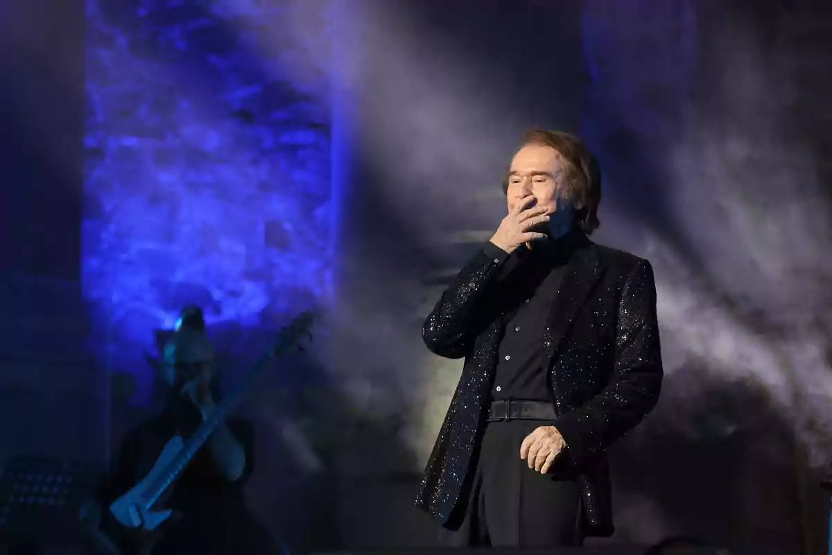 Raphael vestido de negro con chaqueta brillante sobre el escenario llevando una mano a la boca mientras un músico toca el bajo al fondo con luces azules iluminando la escena Raphael vestido de negro con chaqueta brillante sobre el escenario llevando una mano a la boca mientras un músico toca el bajo al fondo con luces azules iluminando la escena