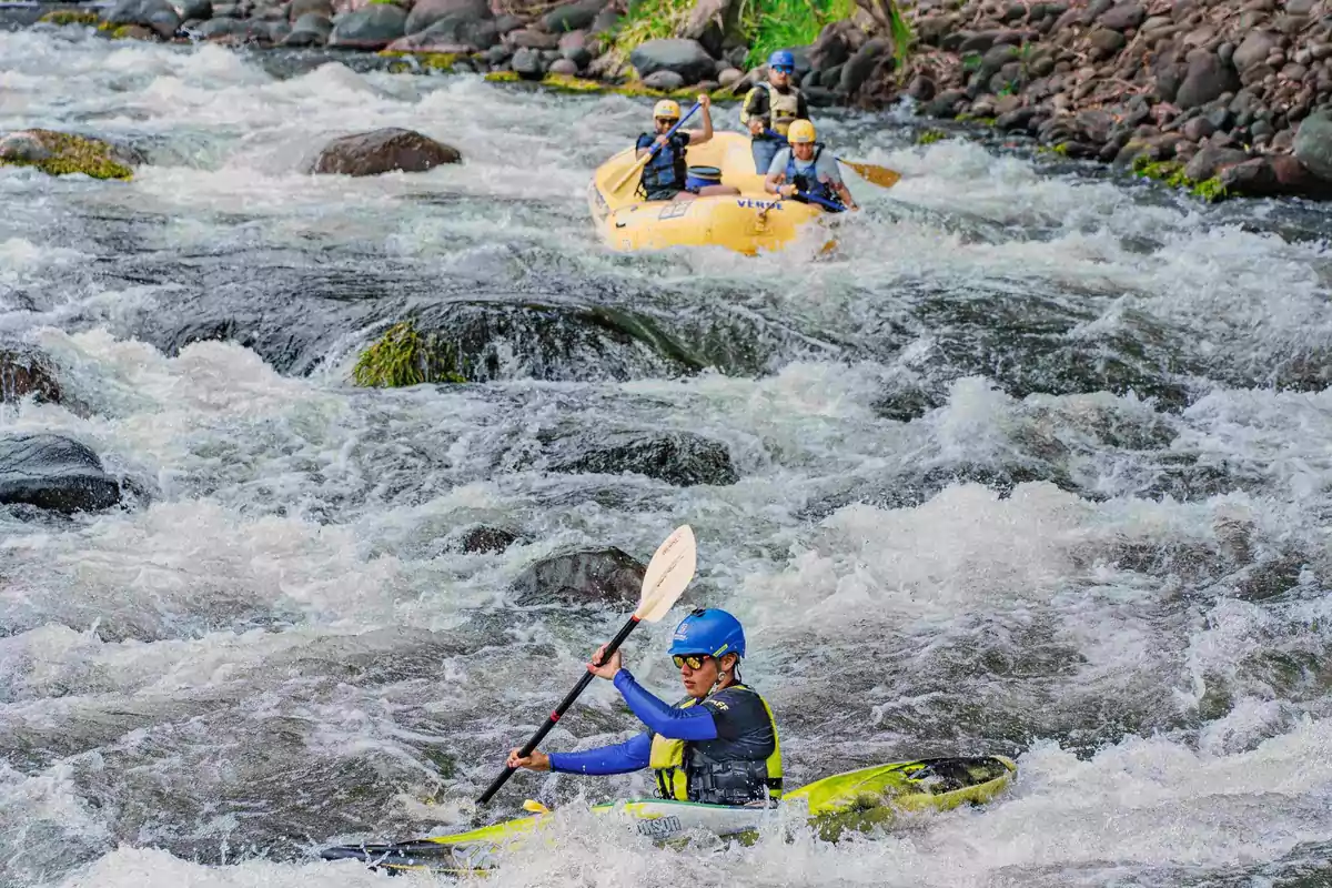 Person in a kayak navigating through rapids while a group in a raft approaches from the background.