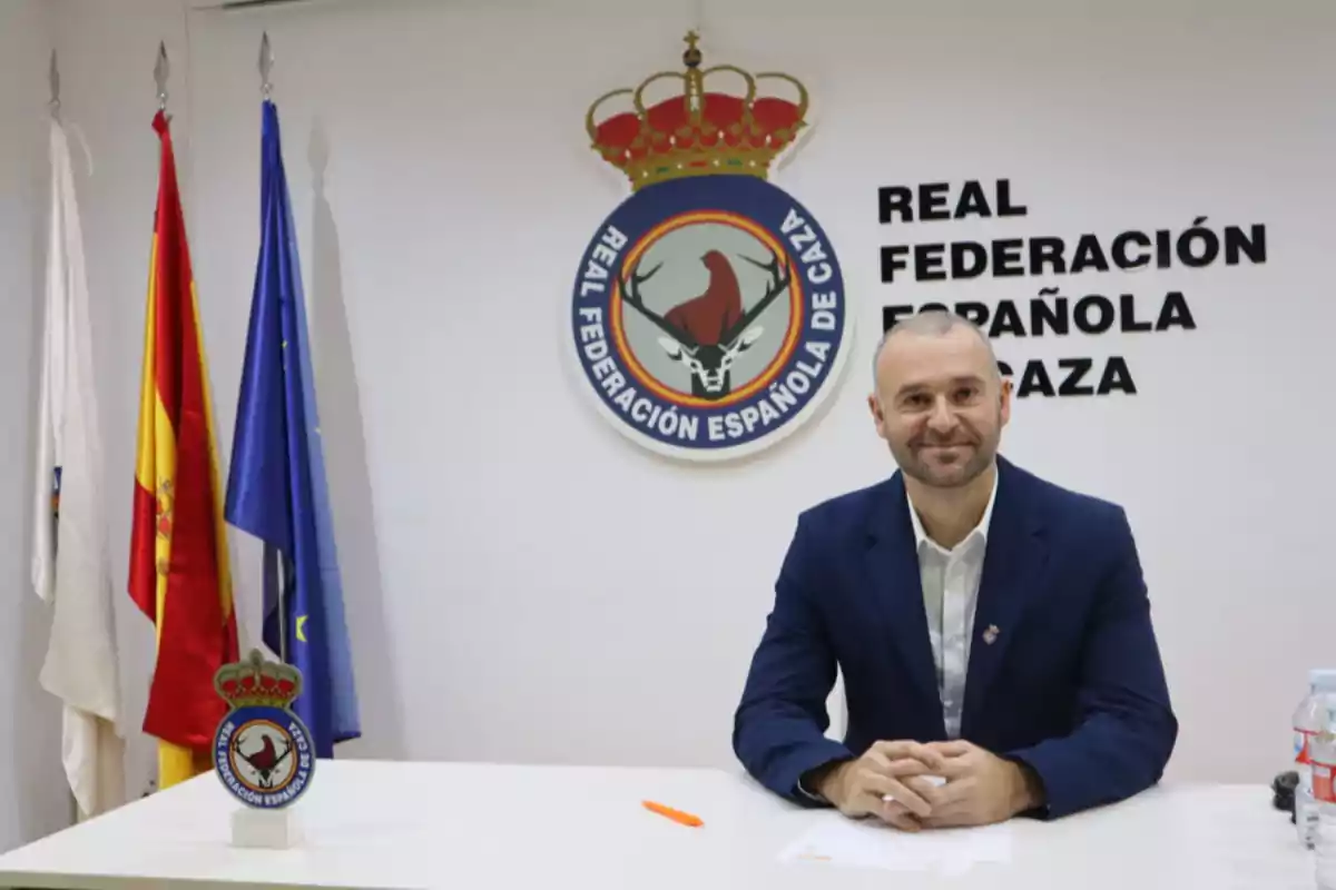 Un hombre sentado en una mesa frente a un logotipo de la Real Federación Española de Caza con varias banderas en el fondo.