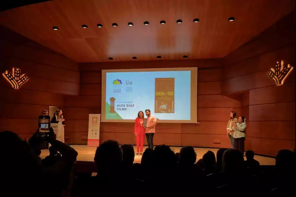 Personas en un escenario de auditorio con una pantalla grande que muestra un cartel de película y un texto que dice "Alex Díaz Films" mientras el público observa.