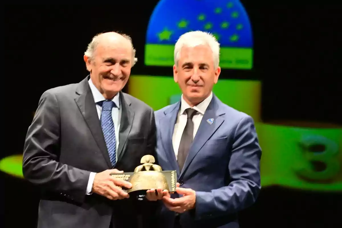 Two men in suits holding a trophy at an event with a dark background.