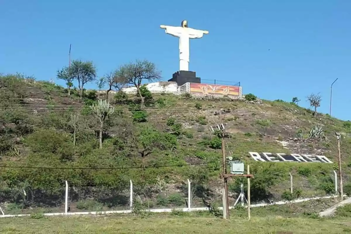 Una estatua de Cristo con los brazos extendidos se encuentra en la cima de una colina rodeada de vegetación y un cielo despejado.