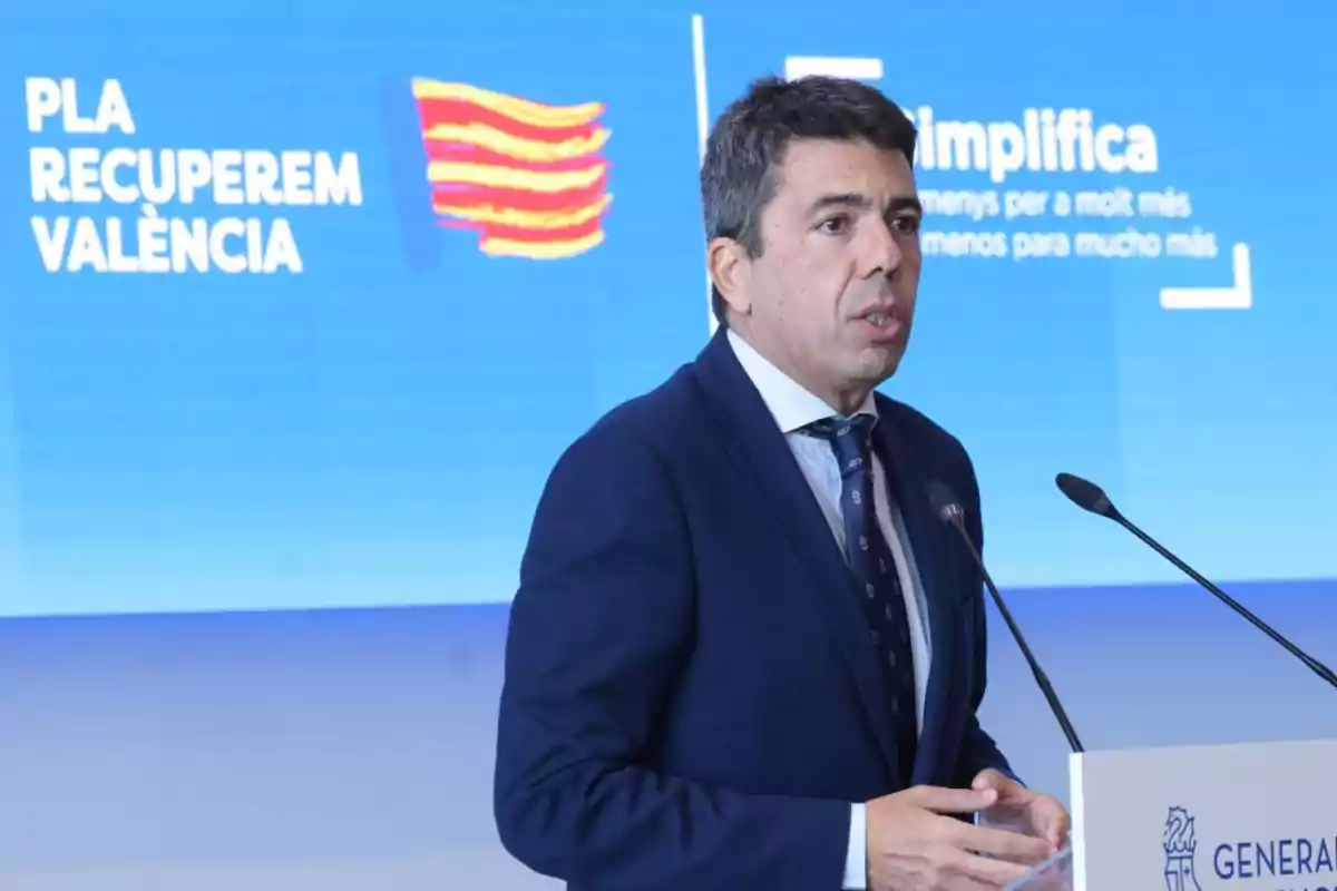 A man in a suit speaks at a podium with a blue background displaying the text "PLA RECUPEREM VALÈNCIA" and "Simplifica".