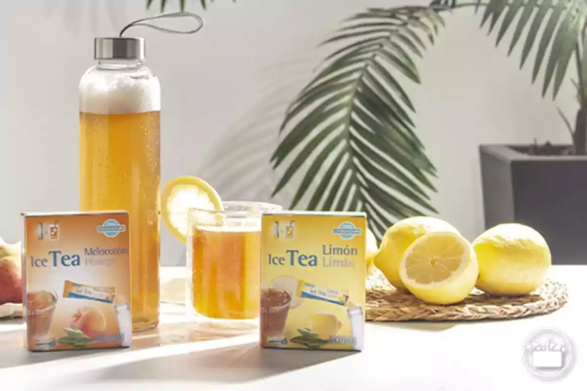 Bebida de té helado en botella y vaso con rodaja de limón junto a cajas de té sabor melocotón y limón y limones frescos sobre una mesa blanca con hojas verdes al fondo Bebida de té helado en botella y vaso con rodaja de limón junto a cajas de té sabor melocotón y limón y limones frescos sobre una mesa blanca con hojas verdes al fondo