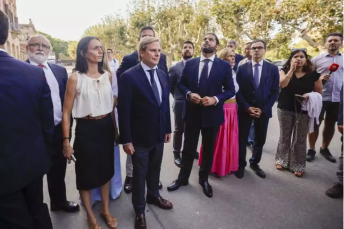 Un grupo de personas de pie al aire libre, algunas vestidas formalmente y otras casualmente, observando algo fuera de la imagen.