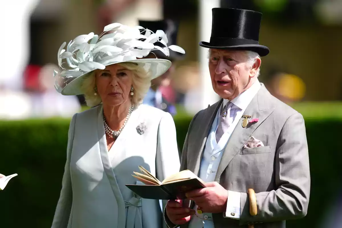 La reina Camila y Carlos III elegantemente vestida asiste a un evento al aire libre, la mujer lleva un sombrero decorativo blanco y el hombre un sombrero de copa negro y sostiene un libro. La reina Camila y Carlos III elegantemente vestida asiste a un evento al aire libre, la mujer lleva un sombrero decorativo blanco y el hombre un sombrero de copa negro y sostiene un libro.