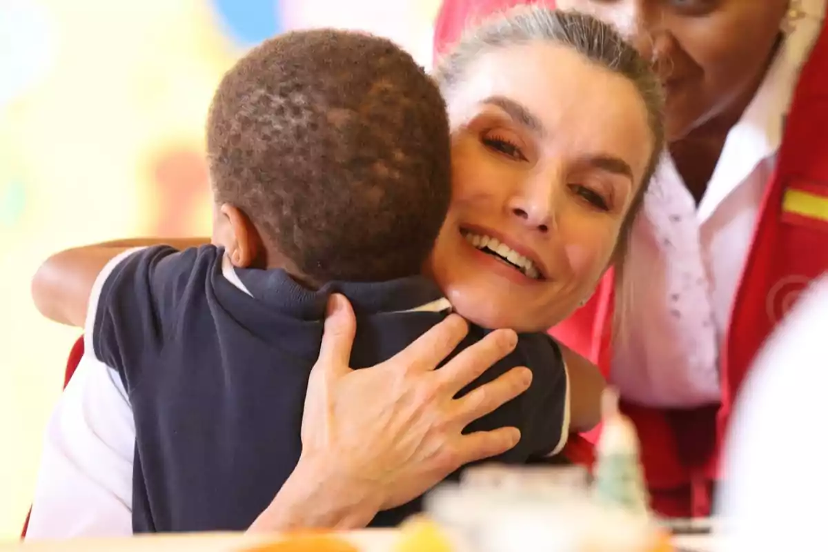 Queen Letizia, smiling, hugs a small child while another person watches in the background. Queen Letizia, smiling, hugs a small child while another person watches in the background.