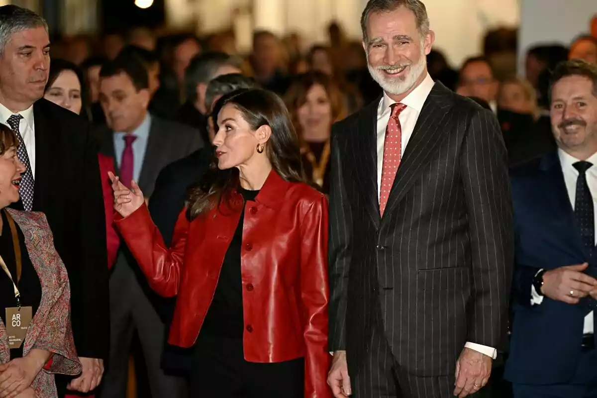 Un grupo de personas vestidas formalmente en un evento, con la reina Letizia de chaqueta roja hablando y Felipe VI con traje a rayas sonriendo. Un grupo de personas vestidas formalmente en un evento, con la reina Letizia de chaqueta roja hablando y Felipe VI con traje a rayas sonriendo.