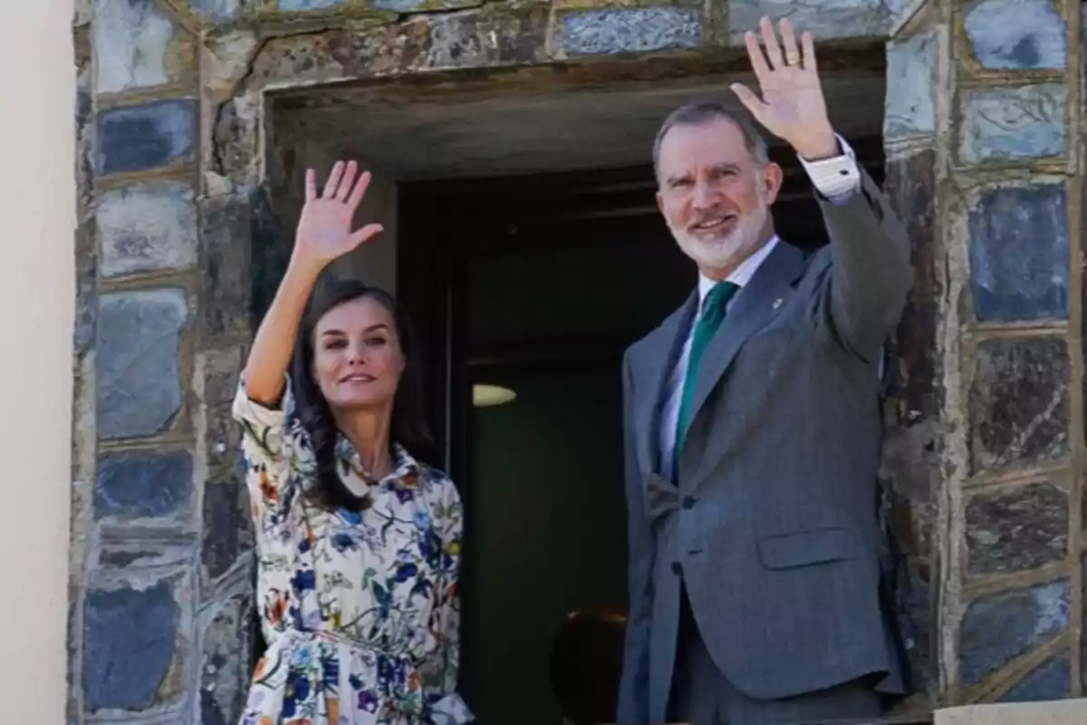 La reina Letizia con vestido floral y Felipe VI con traje gris saludan sonrientes desde una ventana de piedra. La reina Letizia con vestido floral y Felipe VI con traje gris saludan sonrientes desde una ventana de piedra.