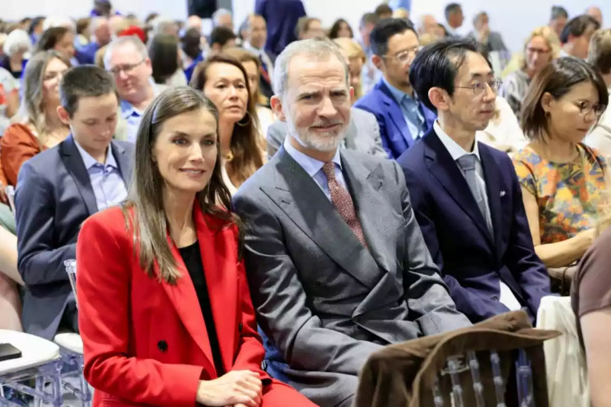 A group of people seated at an event, with a woman in a red suit and a man in a gray suit in the foreground, surrounded by other attendees. A group of people seated at an event, with a woman in a red suit and a man in a gray suit in the foreground, surrounded by other attendees.