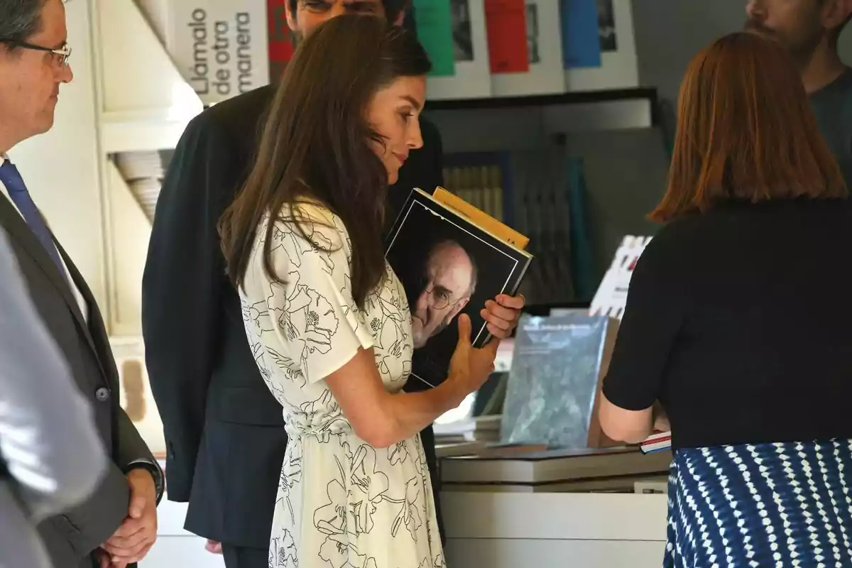 Queen Letizia, wearing a light-colored dress, holds a book with the cover featuring an older man while talking to another person in a bookstore. Queen Letizia, wearing a light-colored dress, holds a book with the cover featuring an older man while talking to another person in a bookstore.