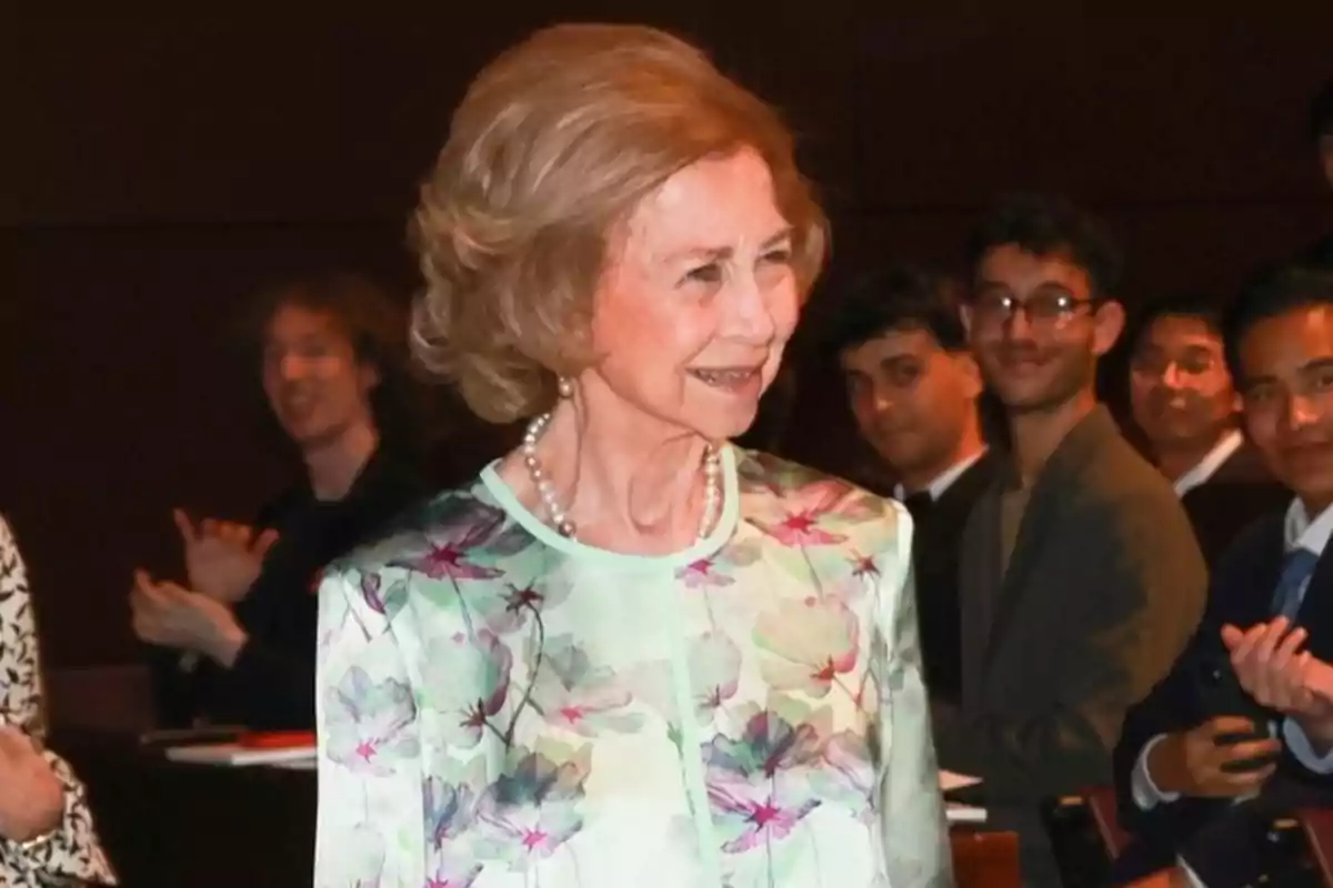 Queen Sofía, wearing a floral dress, smiles while several people watch and applaud her at an event. Queen Sofía, wearing a floral dress, smiles while several people watch and applaud her at an event.