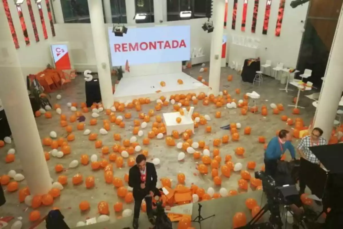 La sede de ciudadanos repleta de globos naranjas y blancos, con una pantalla de fondo que reza "REMONTADA" y con solamente tres personas en su interior