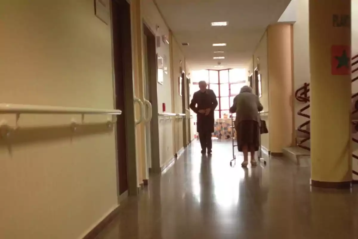 Dos personas mayores caminan por un pasillo iluminado de una residencia o centro de salud, una de ellas usa un andador Dos personas mayores caminan por un pasillo iluminado de una residencia o centro de salud, una de ellas usa un andador