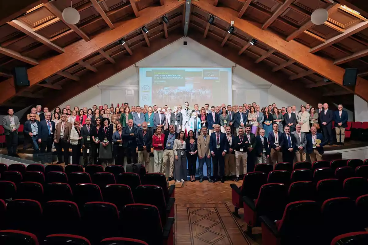 Un grupo grande de personas posando en un auditorio con techo de madera y una pantalla de proyección al fondo.