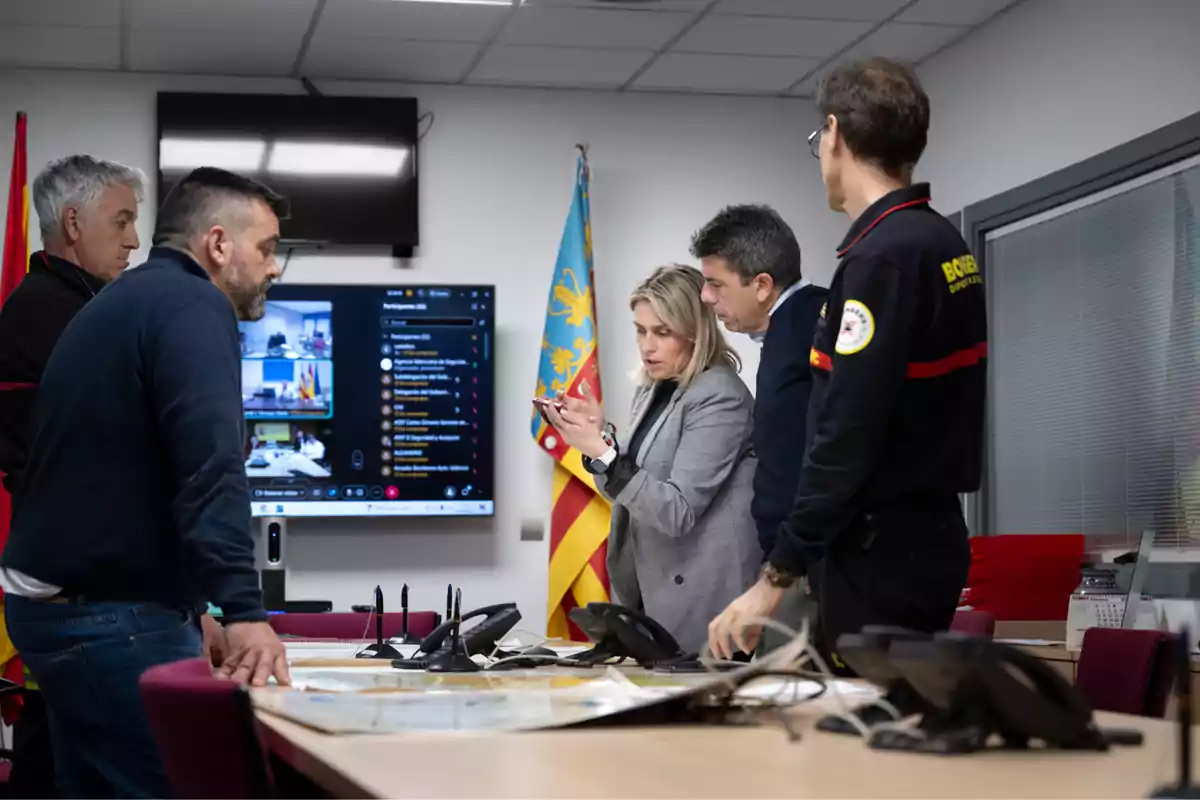 A group of people is in a meeting room looking at a mobile device, with a large screen in the background showing a video conference and flags on the wall.