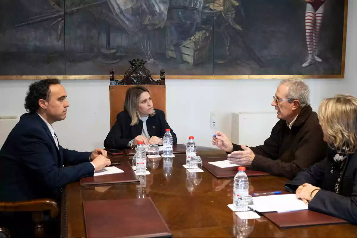 A group of people sitting around a table in a meeting, with water bottles and folders on the table and a large painting on the wall behind them.