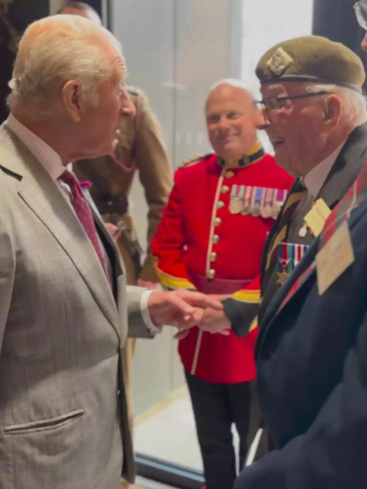 A gray-haired man in a light suit greets and smiles while talking with a military veteran wearing a uniform, beret, and several medals, while another man in a red uniform with medals watches the scene in the background. A gray-haired man in a light suit greets and smiles while talking with a military veteran wearing a uniform, beret, and several medals, while another man in a red uniform with medals watches the scene in the background.