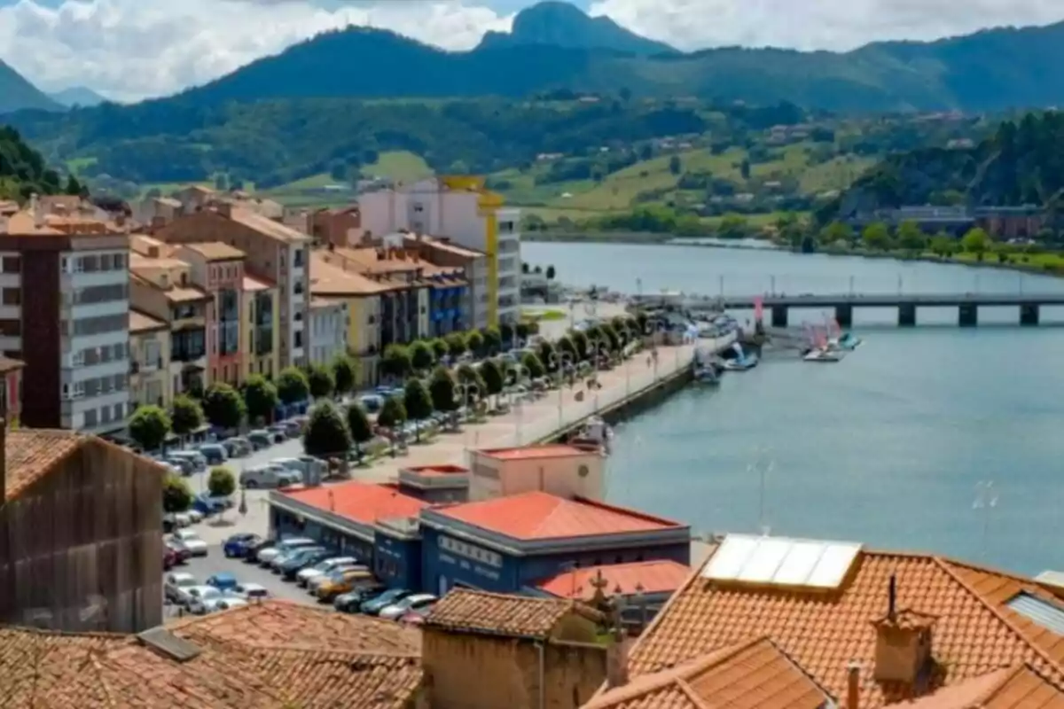 Vista panorámica de una ciudad costera con edificios de colores, un paseo marítimo, un río y montañas al fondo.