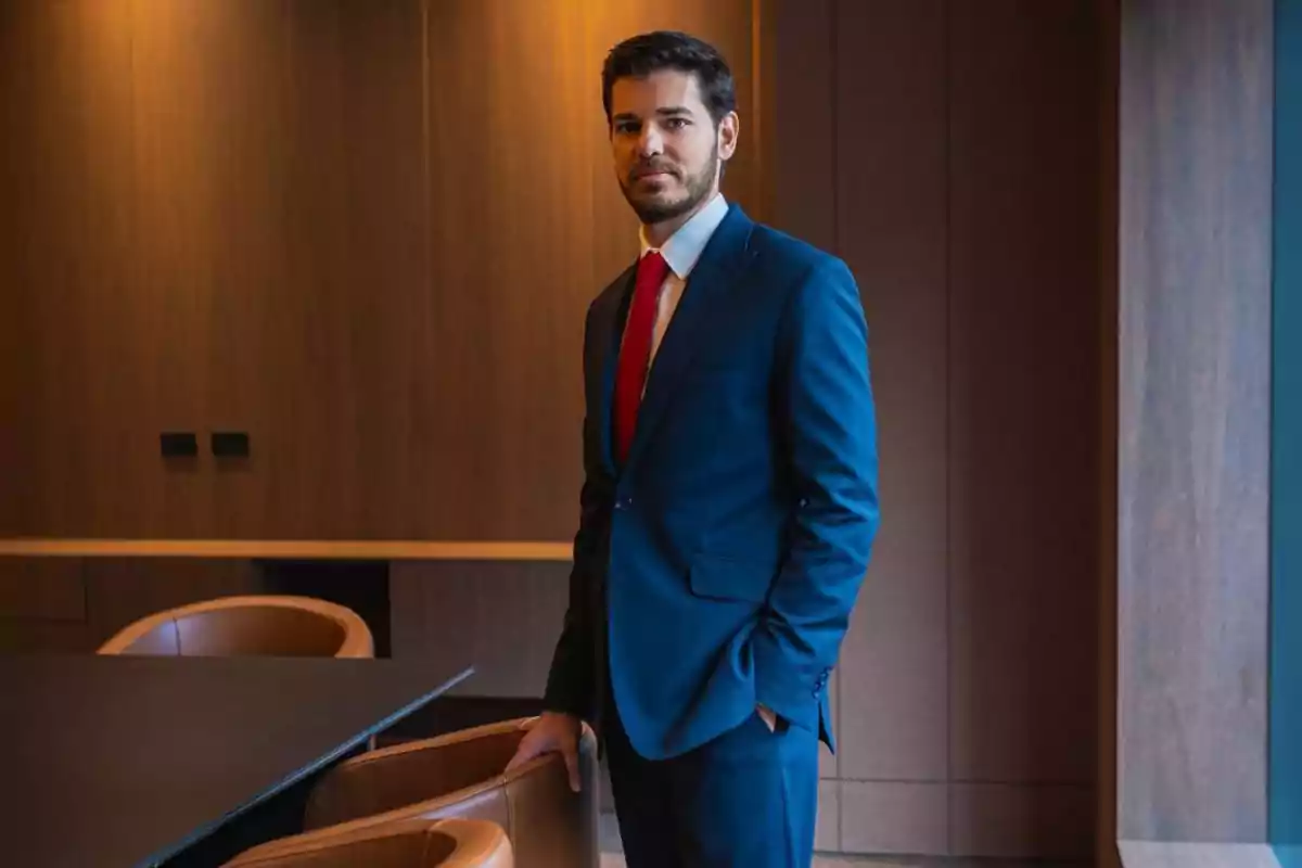 Un hombre con traje azul y corbata roja está de pie junto a una mesa en una sala de reuniones con paredes de madera.