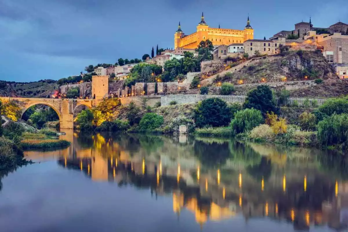 Foto en plano general del Río Tagus en Toledo, España, con luces encendidas que se reflejan en el río junto a las casas y el puente al fondo