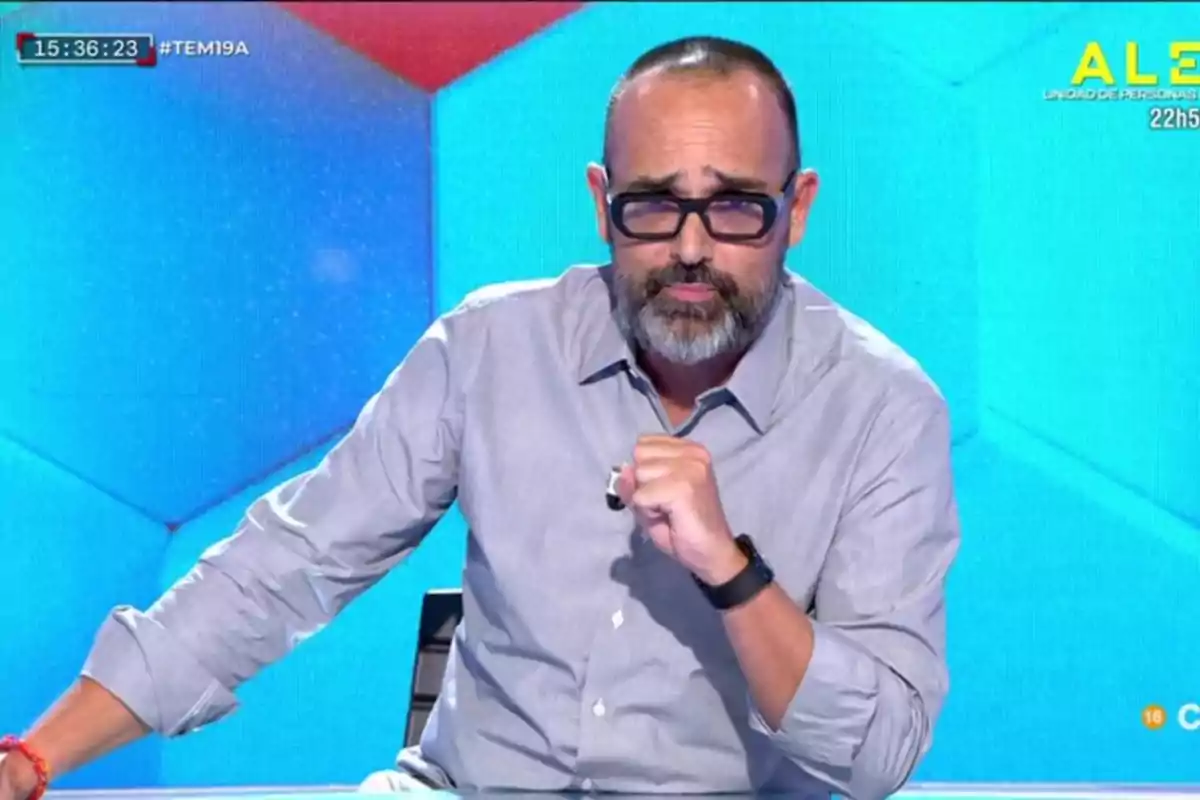 Un hombre con gafas y barba, vestido con una camisa gris, está sentado frente a un fondo azul con patrones geométricos.