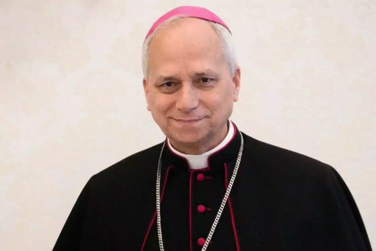 Un hombre con vestimenta eclesiástica y birrete rosa sonríe frente a un fondo claro. Un hombre con vestimenta eclesiástica y birrete rosa sonríe frente a un fondo claro.