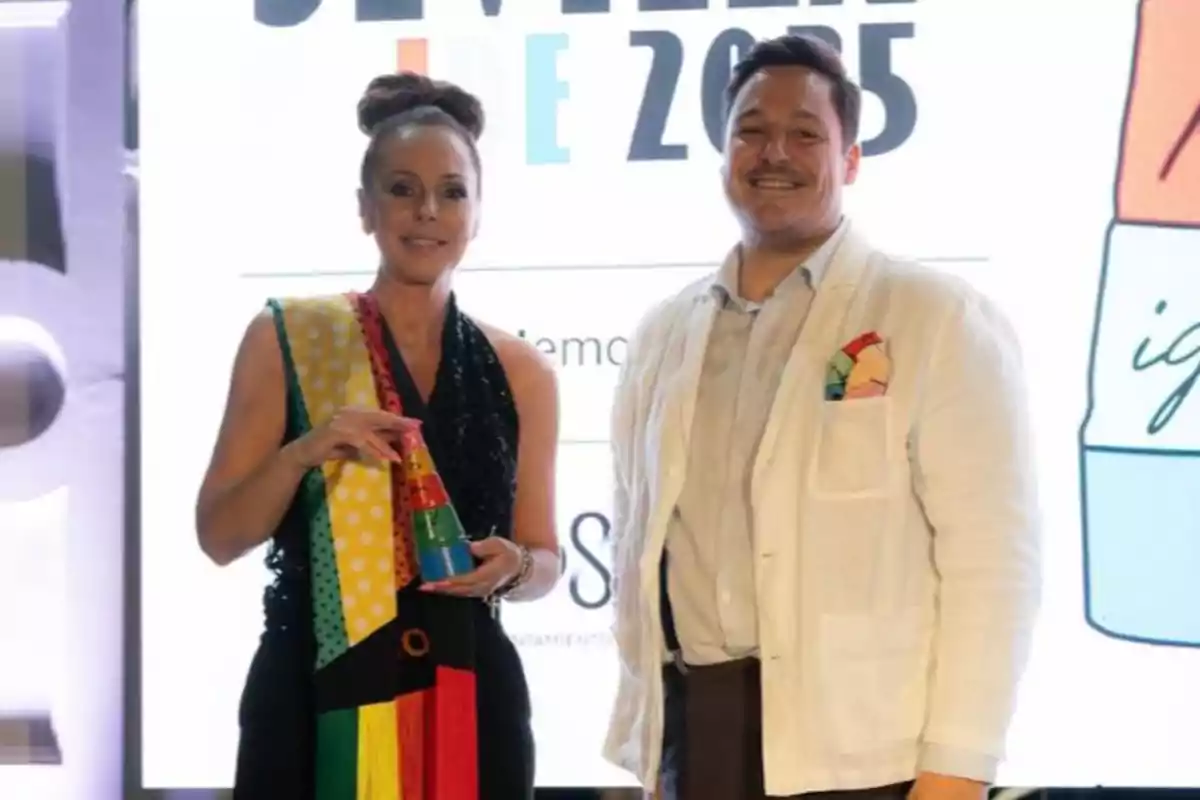 Rocío Carrasco y un hombre posan sonrientes en un evento, una de ellas sostiene un trofeo colorido y ambos llevan prendas con detalles llamativos frente a un fondo iluminado con texto. Rocío Carrasco y un hombre posan sonrientes en un evento, una de ellas sostiene un trofeo colorido y ambos llevan prendas con detalles llamativos frente a un fondo iluminado con texto.