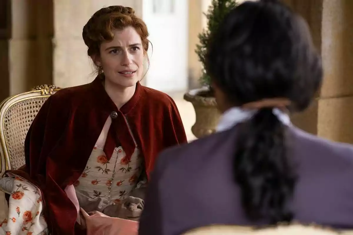 Rocío Suárez de Pugar con vestido de época y capa roja está sentada en una silla, conversando Marco Pernas de espaldas en un entorno elegante en Valle Salvaje. Rocío Suárez de Pugar con vestido de época y capa roja está sentada en una silla, conversando Marco Pernas de espaldas en un entorno elegante en Valle Salvaje.