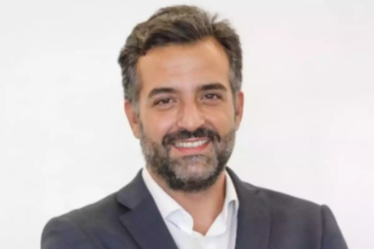 Hombre de cabello y barba oscura con saco azul y camisa blanca sonriendo frente a un fondo claro