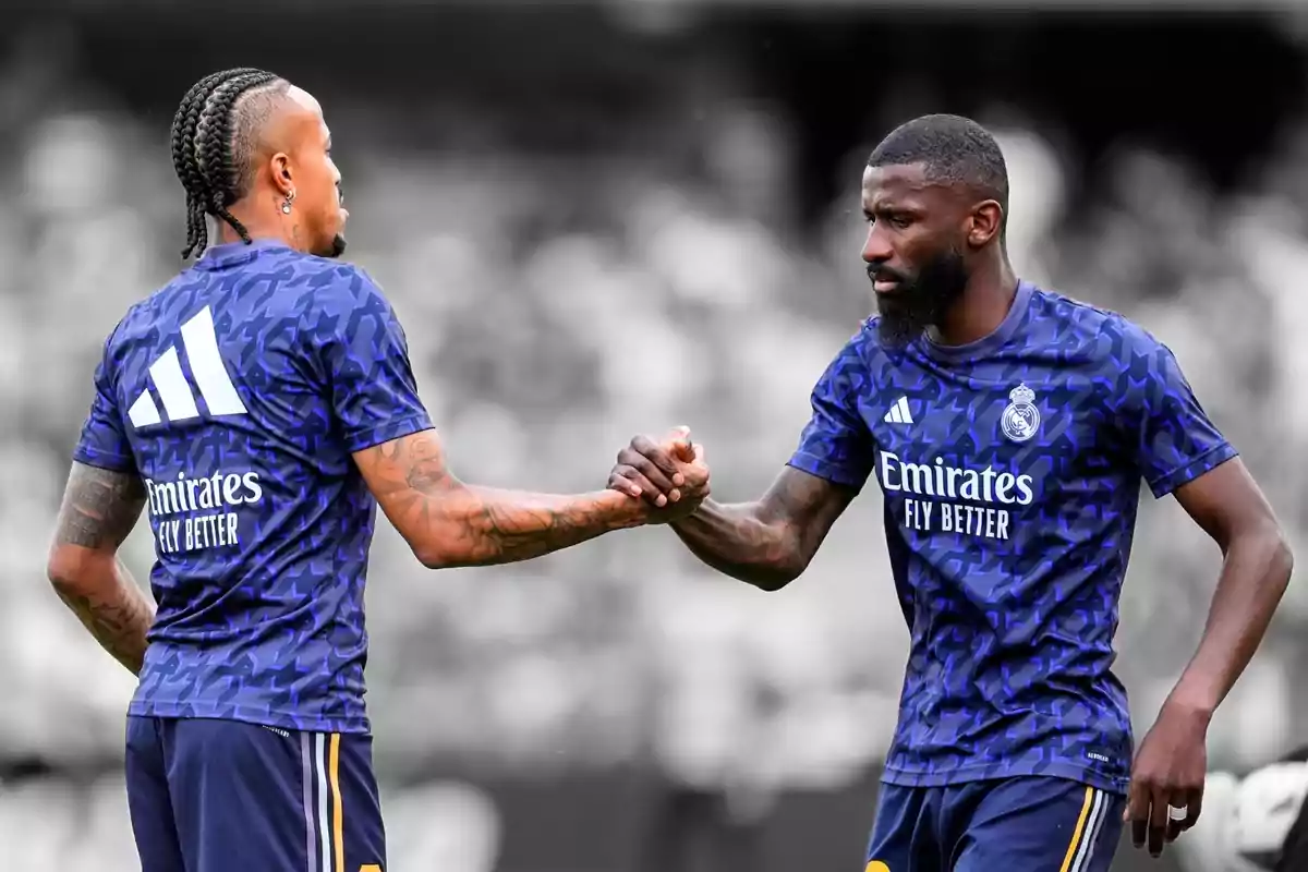 Dos jugadores de fútbol con uniformes de entrenamiento del Real Madrid se dan la mano en el campo.