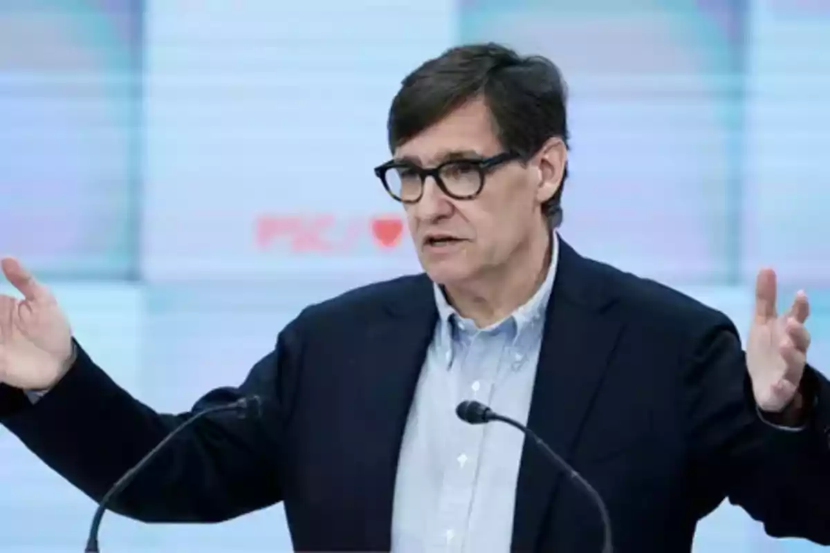 Hombre con gafas y chaqueta oscura hablando en una conferencia con los brazos extendidos frente a dos micrófonos Hombre con gafas y chaqueta oscura hablando en una conferencia con los brazos extendidos frente a dos micrófonos