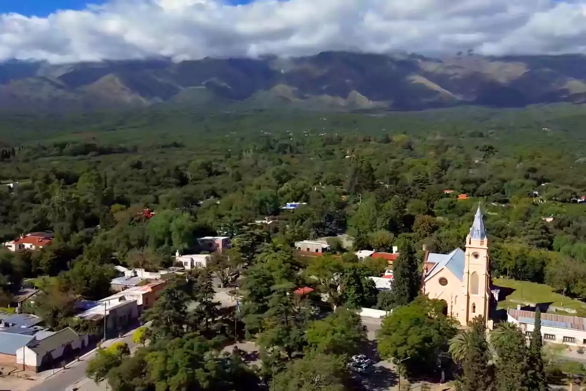 Vista aérea de un pueblo rodeado de vegetación con una iglesia de torre alta en primer plano y montañas cubiertas de nubes al fondo Vista aérea de un pueblo rodeado de vegetación con una iglesia de torre alta en primer plano y montañas cubiertas de nubes al fondo