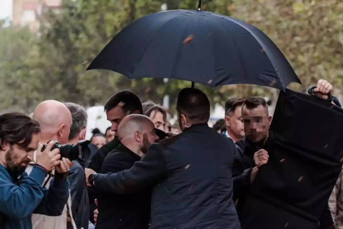 Un grupo de personas rodea a un individuo bajo un paraguas negro mientras un fotógrafo captura el momento. Un grupo de personas rodea a un individuo bajo un paraguas negro mientras un fotógrafo captura el momento.