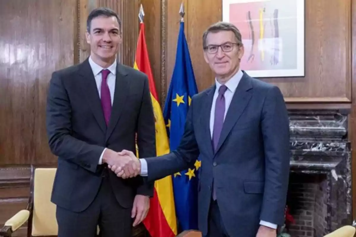 Dos hombres de traje y corbata se dan la mano frente a las banderas de España y la Unión Europea en una oficina con paredes de madera.