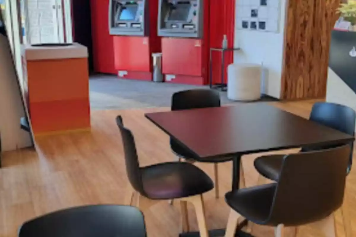 Una mesa con cuatro sillas negras en un área de espera con dos cajeros automáticos al fondo. Una mesa con cuatro sillas negras en un área de espera con dos cajeros automáticos al fondo.