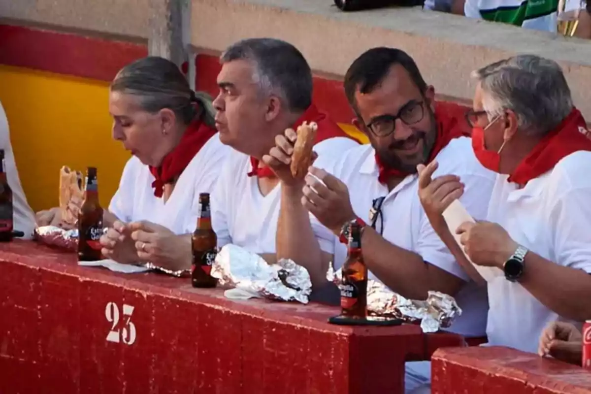 Personas vestidas de blanco con pañuelos rojos en el cuello, sentadas en una grada, comiendo bocadillos y bebiendo cerveza.