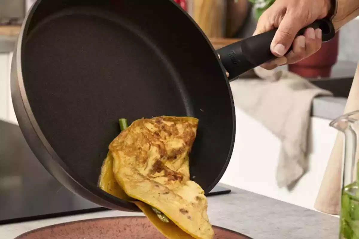 Mano volcando una tortilla de espárragos de una sartén sobre un plato, en la cocina Mano volcando una tortilla de espárragos de una sartén sobre un plato, en la cocina