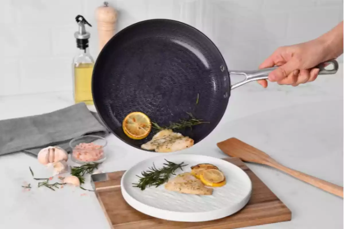 Una sartén negra con mango de metal vierte filetes de pescado con rodajas de limón y romero en un plato blanco sobre una tabla de madera, rodeado de ingredientes como ajo, sal rosada y aceite de oliva. Una sartén negra con mango de metal vierte filetes de pescado con rodajas de limón y romero en un plato blanco sobre una tabla de madera, rodeado de ingredientes como ajo, sal rosada y aceite de oliva.