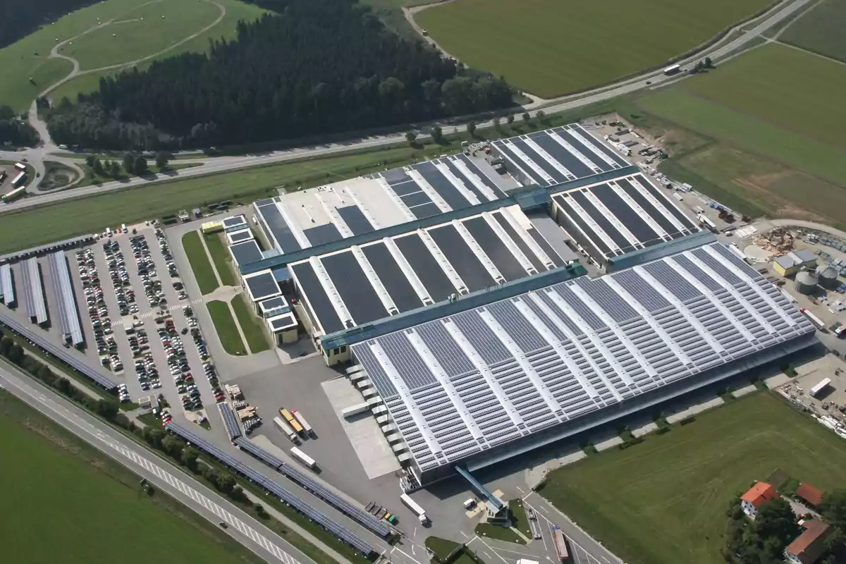 Vista aérea de una gran instalación industrial con techos cubiertos de paneles solares rodeada de áreas verdes y estacionamientos.