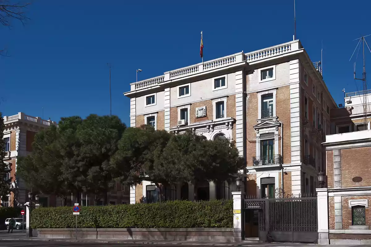 Edificio de ladrillo con detalles blancos rodeado de árboles y una reja en la entrada bajo un cielo azul claro.