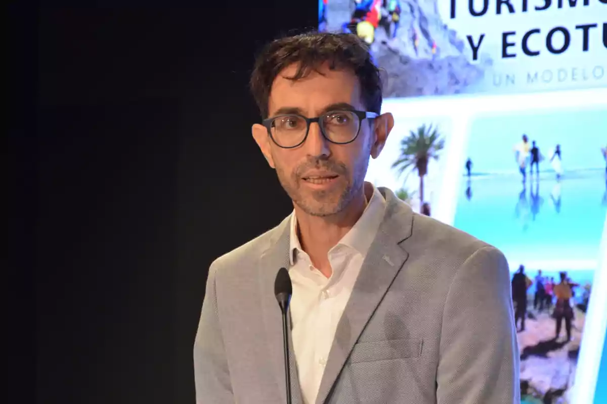 Hombre con gafas y saco gris hablando en un micrófono con un fondo de imágenes relacionadas con turismo y ecoturismo.